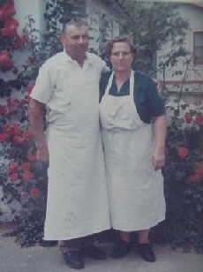 Couple in white aprons stands together outside a home adorned with blooming roses, showcasing their shared culinary passion and warmth.