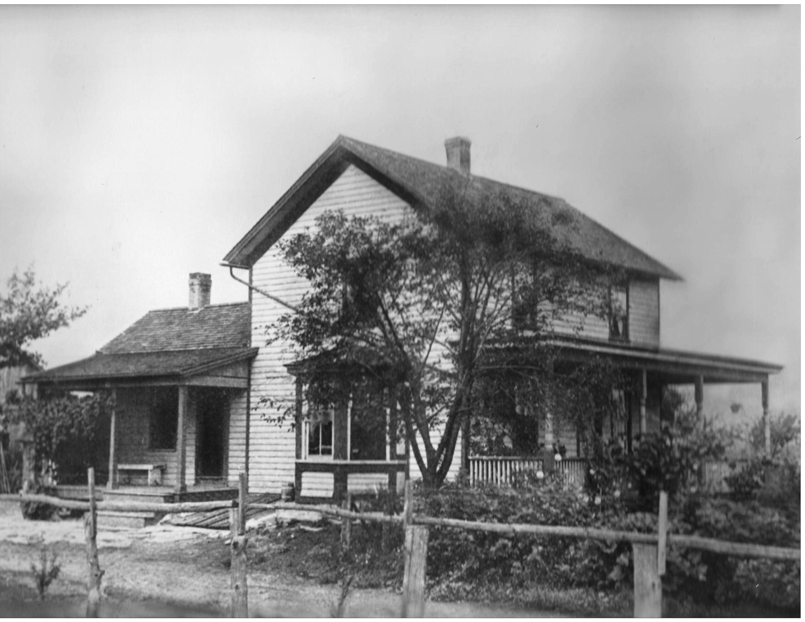 Historic two-story farmhouse with a porch, surrounded by trees and a rustic fence. The image captures early 20th-century rural architecture.