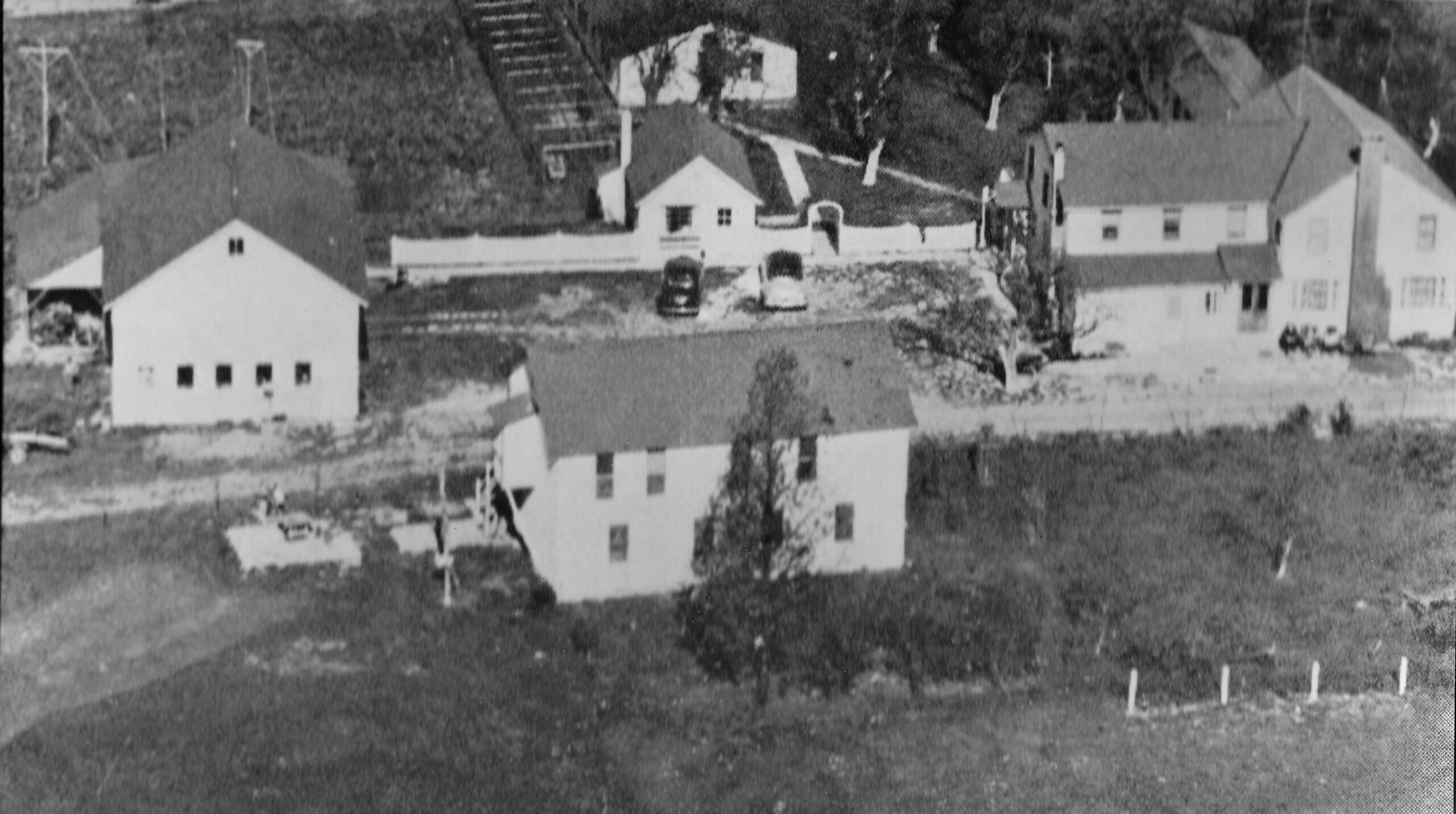 Aerial view of multiple rural buildings, including houses and a barn, surrounded by grassland and a gravel drive, illustrating a quiet countryside setting.