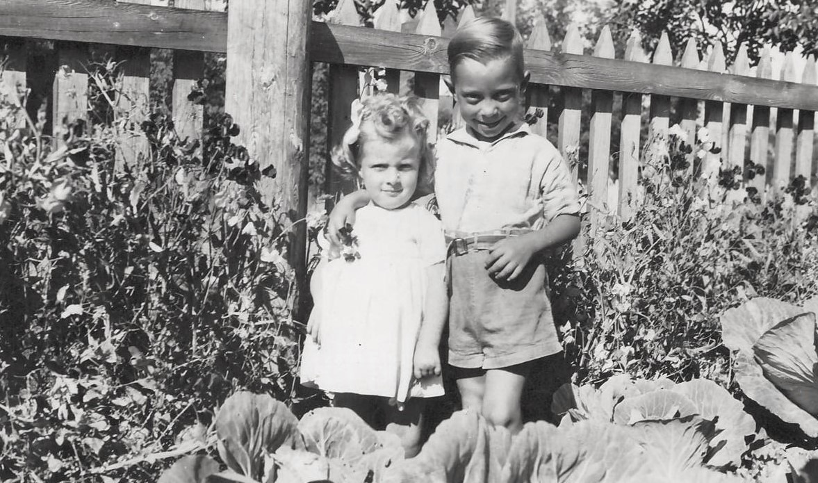 Two children stand together in a garden, smiling. The girl wears a white dress with a bow, while the boy is in shorts and a shirt. Cabbage plants and flowers surround them.
