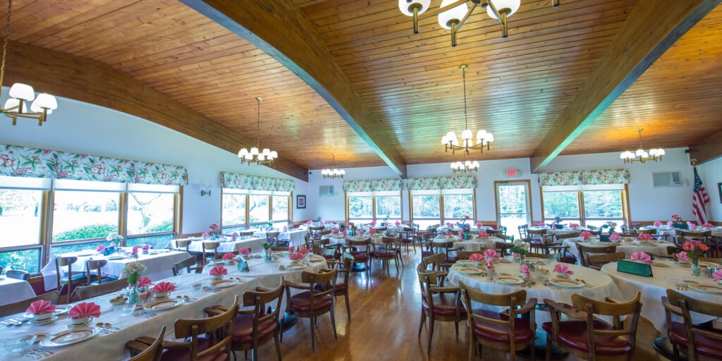 A spacious banquet hall with wood-paneled ceilings and large windows. Tables are set with white tablecloths, pink napkins, and floral decor, creating an elegant atmosphere.