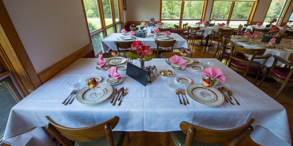 Elegant dining room with white-clothed tables set for guests. Pink napkins, floral centerpieces, and sunlight from large windows create a warm ambiance.