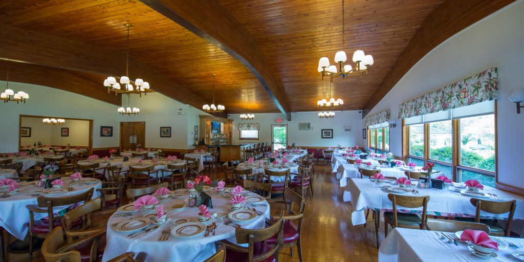 Spacious dining room with wooden ceiling, warmly lit by chandeliers. Tables are elegantly set with white tablecloths and pink napkins, creating an inviting atmosphere.
