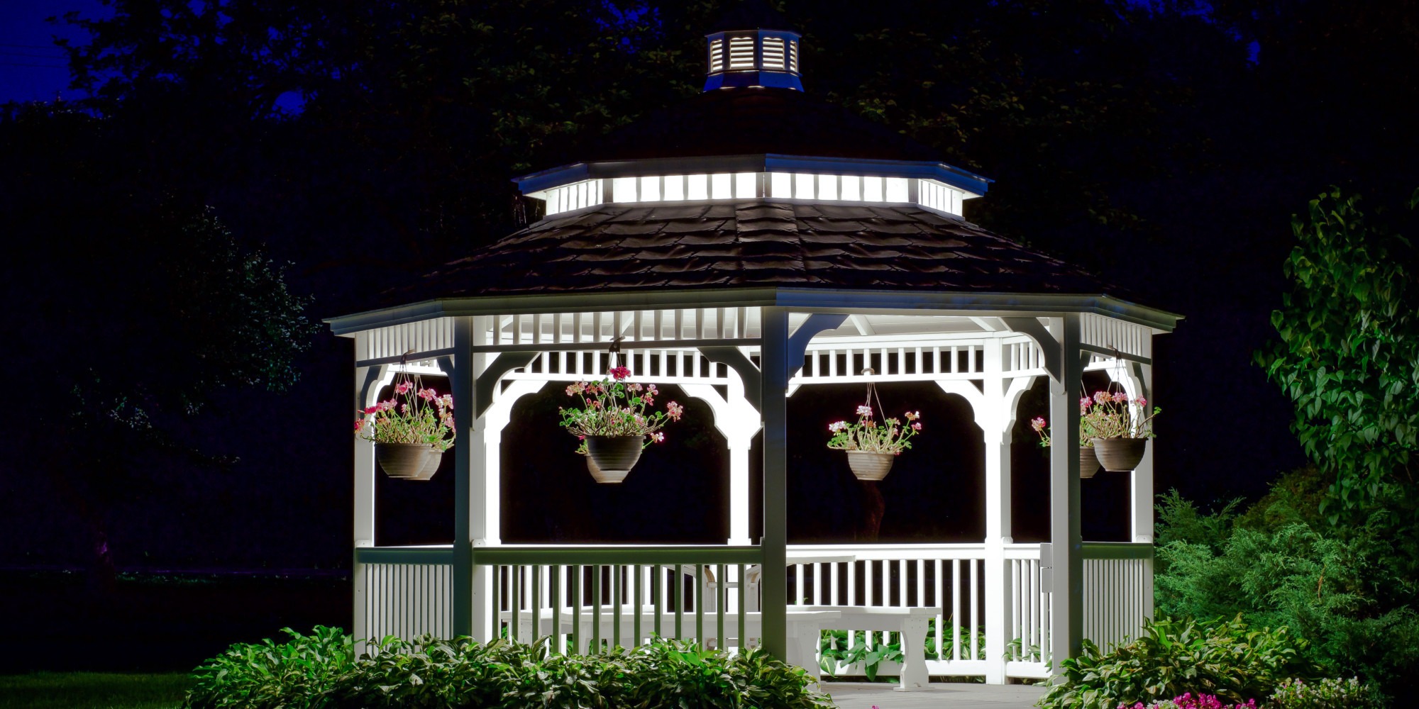 A beautifully illuminated gazebo at night, adorned with hanging flower pots, creating a serene outdoor atmosphere in a garden setting.