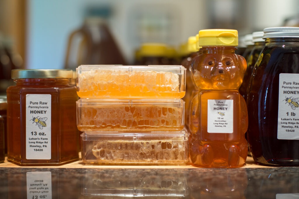 Various honey containers, including jars and a bear-shaped bottle, are displayed. Honeycomb in plastic boxes in the center adds a natural touch.