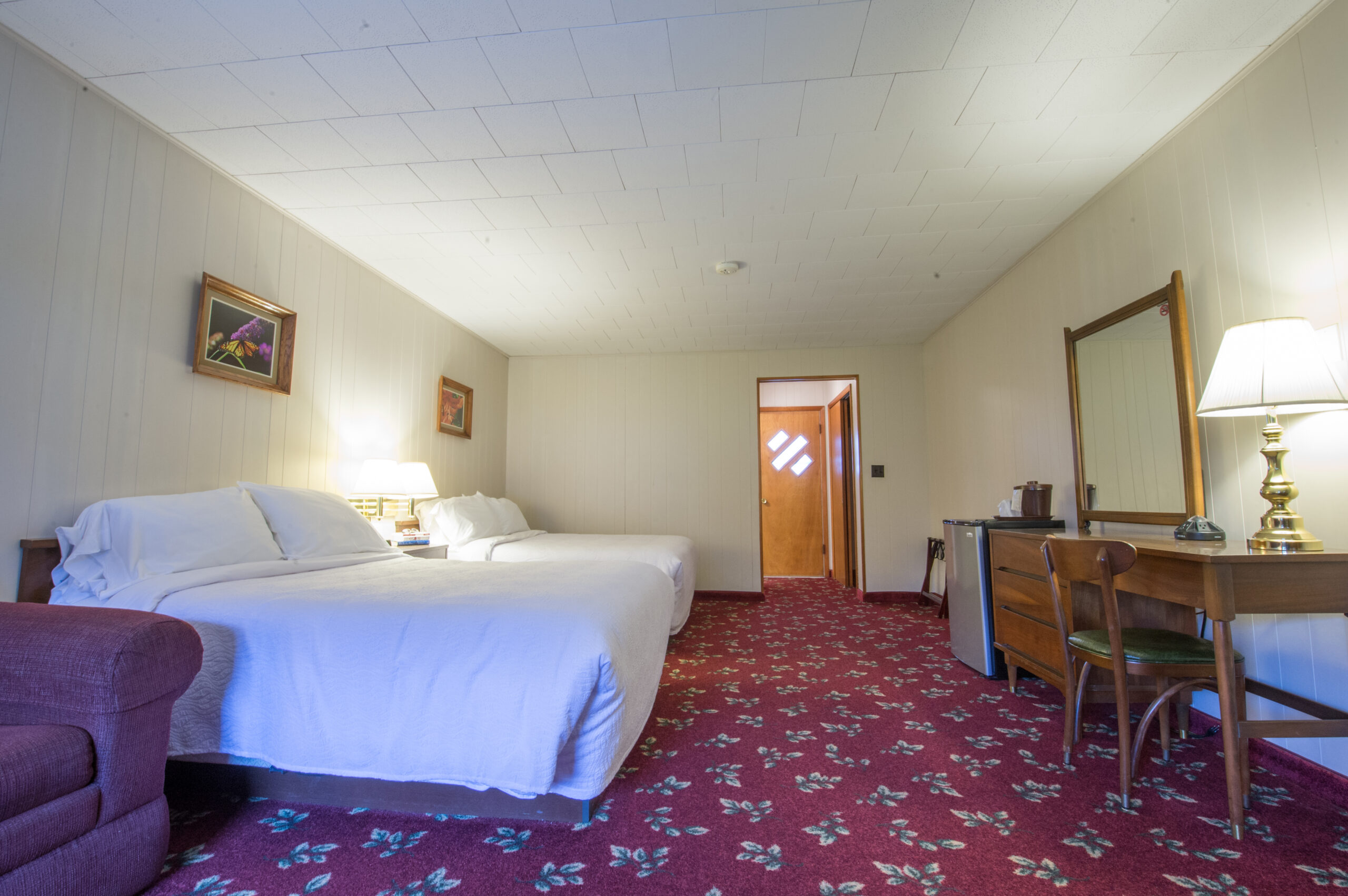 Spacious hotel room with two beds, a sitting area, and a wooden desk. Decorated with floral carpets and framed art, featuring a windowed door.